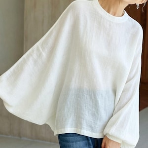May include: A white, long-sleeved blouse with a round neckline and loose, flowing sleeves. The fabric appears to be a lightweight linen or similar material. The blouse is a solid, off-white color.