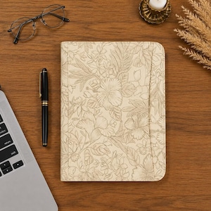May include: A beige tablet or notebook cover featuring a detailed line-art floral pattern with hibiscus and tropical leaves. The cover is positioned next to a black fountain pen, a pair of thin-rimmed glasses, and a partial view of a laptop.