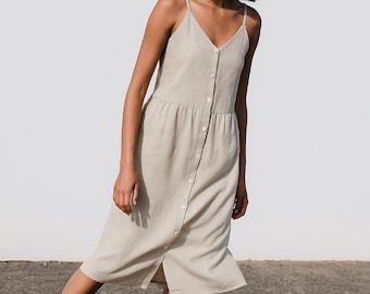 May include: A light beige, button-down midi dress with spaghetti straps. The dress features a V-neckline, a fitted bodice, and a flowing skirt with a front slit. The dress is made of a lightweight fabric, perfect for warm weather.