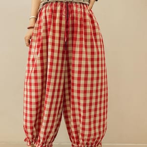 May include: Loose-fitting trousers featuring a red and white gingham check pattern. These pants have an elasticized waistband with a drawstring tie and gathered, ruffled cuffs at the ankles, creating a voluminous silhouette.