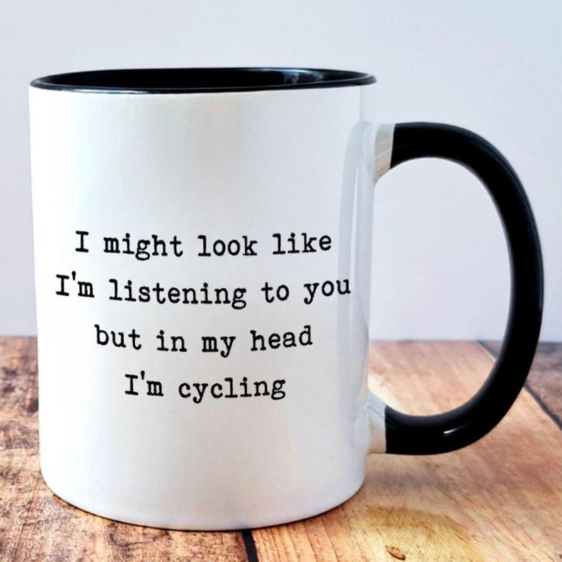 May include: White ceramic mug with a black rim and handle. The mug features the text "I might look like I'm listening to you but in my head I'm cycling" in black font. The mug is on a wooden surface.