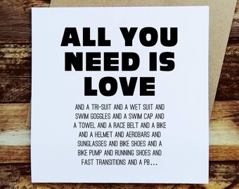 May include: A white greeting card with the text "ALL YOU NEED IS LOVE" in bold black letters. Below, smaller text lists triathlon gear: a tri-suit, wetsuit, goggles, swim cap, towel, race belt, bike, helmet, aerobars, sunglasses, bike shoes, pump, running shoes, and fast transitions.
