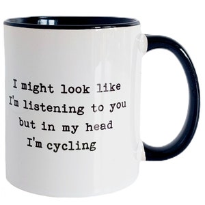 May include: White ceramic mug with a black interior and handle. The mug features the text "I might look like I'm listening to you but in my head I'm cycling" in black, sans-serif font.