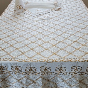 May include: Cream-colored tablecloth with a diamond pattern and gold embroidered details. The tablecloth is on a table with a matching napkin holder and napkins. The border features a repeating floral design.