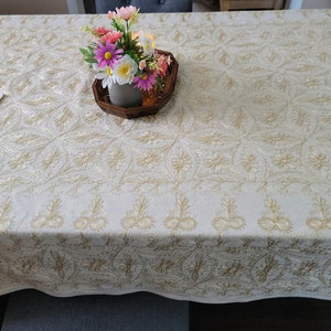 Pode incluir: Toalha creme com bordados dourados intrincados. O padrão floral e de folhas cobre toda a superfície. Uma toalha de mesa a condizer está à esquerda. Uma pequena bandeja de madeira com flores está no centro.
