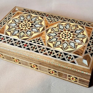 Op de afbeelding: Rechthoekige houten doos met ingelegde geometrische sterpatronen in bruin, beige en crème. De doos heeft decoratieve randen met kleine kleurrijke accenten. Een handgemaakte decoratieve opbergdoos.