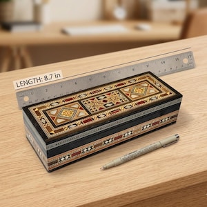 May include: A rectangular wooden box featuring intricate geometric marquetry in shades of brown, cream, and black. A metal ruler placed behind the box indicates a length of 8.7 inches. A grey pen lies on the surface in front of the box.
