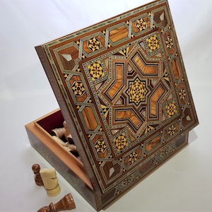 May include: An open wooden chess set box with a detailed geometric inlaid design. The box features a starburst pattern in shades of brown, beige, and yellow. The interior is lined with red fabric, and chess pieces are visible inside.
