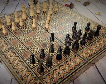 May include: A detailed, wooden chess set with black and cream-colored pieces arranged on a decorative board. The board features inlaid patterns in shades of brown, gold, and cream. The chess pieces are in the starting positions.