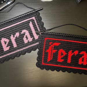 May include: Two crocheted wall hangings with the word "Feral" spelled out in pink and red yarn on a black background.