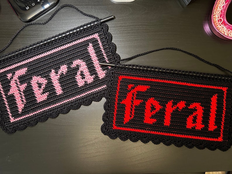 May include: Two crocheted wall hangings with the word "Feral" spelled out in pink and red yarn on a black background.