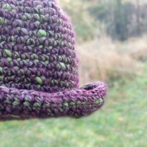 May include: A close-up of a crocheted hat with a purple and green color scheme. The hat is made with a textured yarn and has a wide brim.