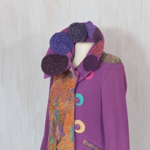 May include: A purple jacket with a colorful crocheted collar and a patterned fabric scarf. The jacket has a variety of colorful crocheted circles sewn on the front.