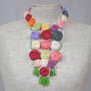 Pu&ograve; includere: Una collana floreale colorata realizzata con rose di stoffa e perline verdi. La collana &egrave; esposta su un manichino bianco.
