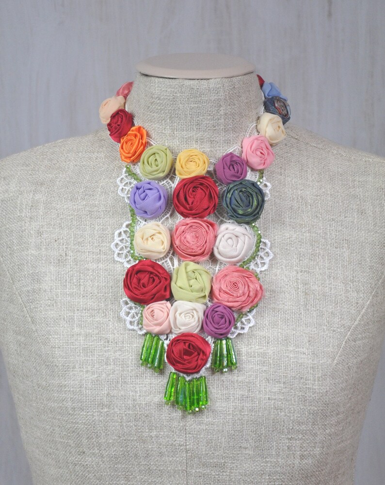 Pu&ograve; includere: Una collana floreale colorata realizzata con rose di stoffa e perline verdi. La collana &egrave; esposta su un manichino bianco.