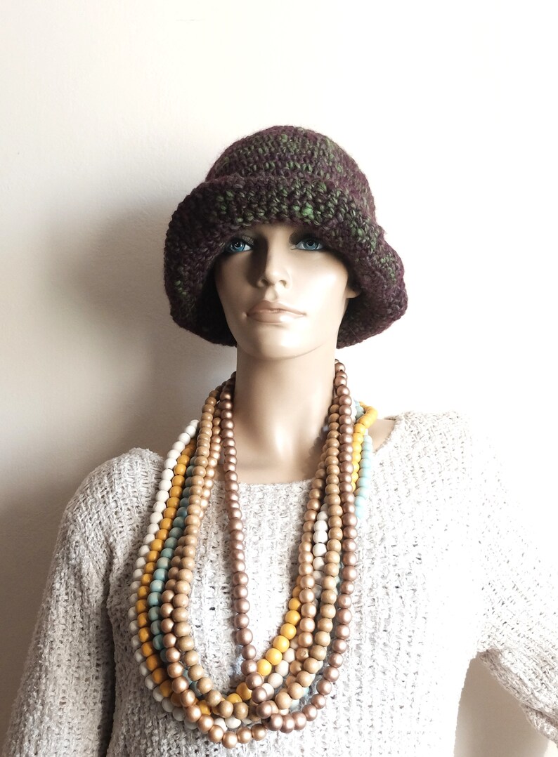 May include: A mannequin wearing a brown and green crocheted hat and a multi-strand necklace of wooden beads in shades of brown, white, and yellow.