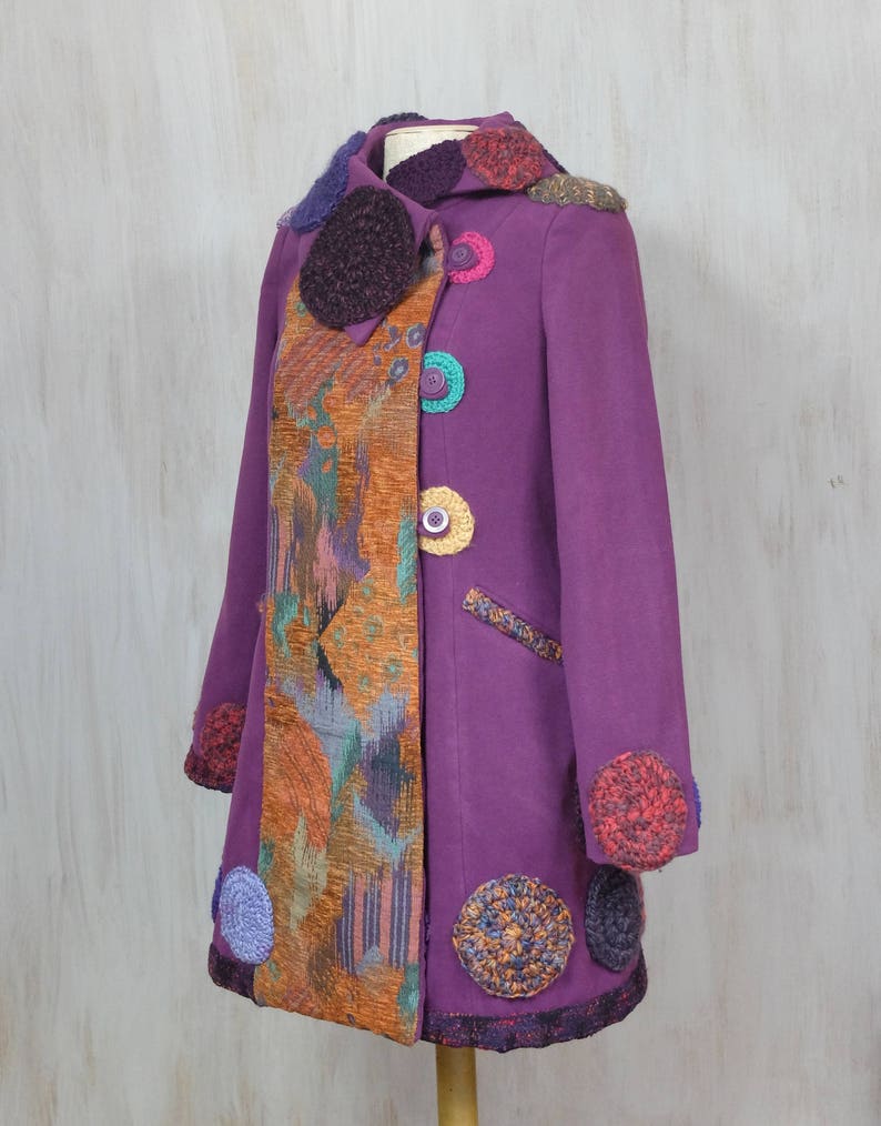 May include: A purple coat with a colorful crocheted trim and buttons. The coat has a patchwork design on one side and a solid color on the other.