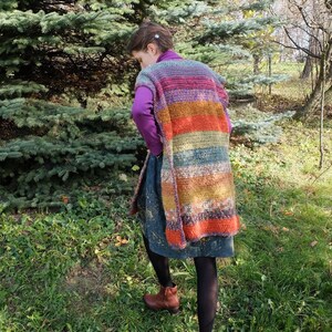May include: A long, multi-colored crochet vest with stripes of purple, orange, brown, and green. The vest is sleeveless and has a loose fit.