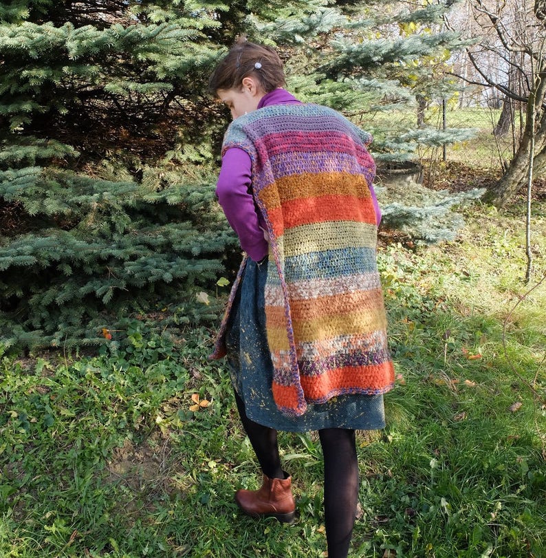 May include: A long, multi-colored crochet vest with stripes of purple, orange, brown, and green. The vest is sleeveless and has a loose fit.