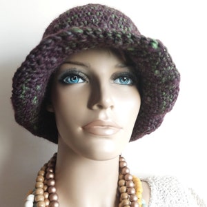 May include: A crocheted hat with a wide brim in shades of purple and green. The hat is worn by a mannequin with a necklace of wooden beads in shades of brown, yellow, and blue.