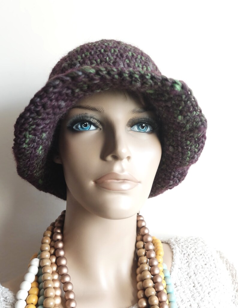 May include: A crocheted hat with a wide brim in shades of purple and green. The hat is worn by a mannequin with a necklace of wooden beads in shades of brown, yellow, and blue.