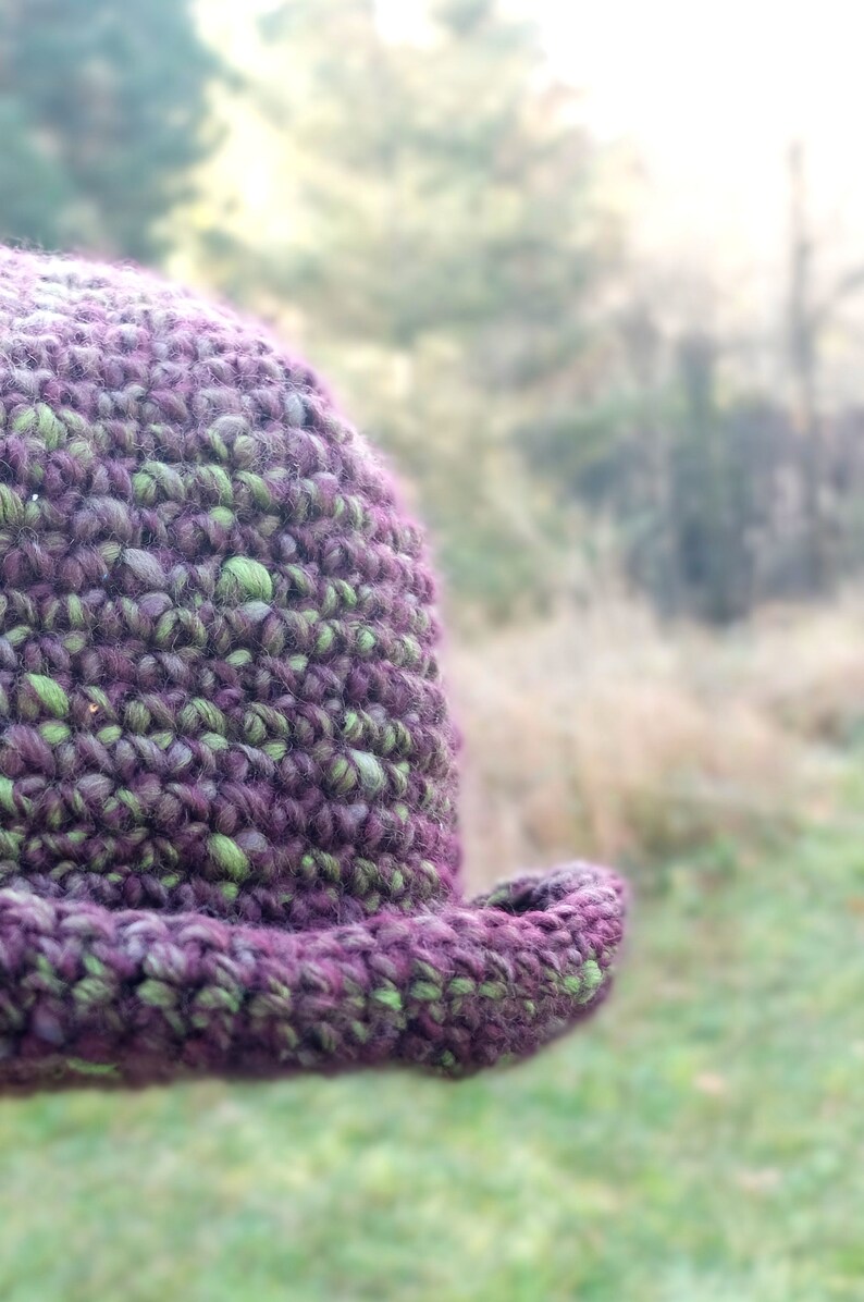May include: A close-up of a crocheted hat with a purple and green color scheme. The hat is made with a textured yarn and has a wide brim.