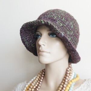 May include: A purple and green crocheted cloche hat with a wide brim. The hat is worn by a mannequin with a multi-colored beaded necklace.