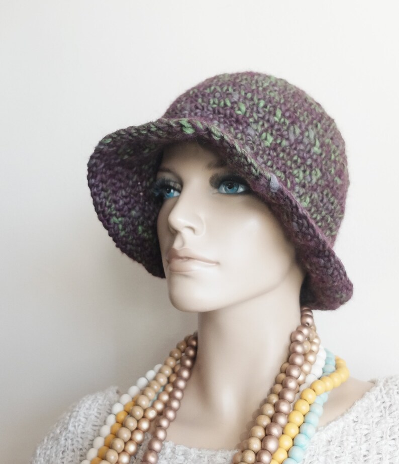 May include: A purple and green crocheted cloche hat with a wide brim. The hat is worn by a mannequin with a multi-colored beaded necklace.