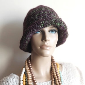 May include: A purple and green crocheted cloche hat with a wide brim. The hat is worn by a mannequin with a multi-colored beaded necklace.