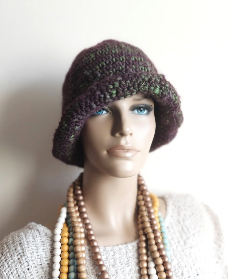 May include: A purple and green crocheted cloche hat with a wide brim. The hat is worn by a mannequin with a multi-colored beaded necklace.