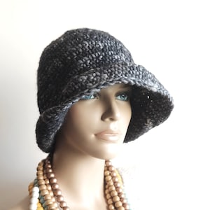 May include: A gray and black crocheted cloche hat with a wide brim. The hat is worn by a mannequin with a long necklace of wooden beads in various shades of brown, white, and blue.