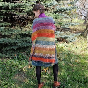 May include: A long, multi-colored crochet cardigan with a striped pattern in shades of purple, orange, red, green, brown, and blue. The cardigan is worn over a dark blue dress.