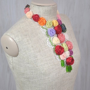 Pu&ograve; includere: Una collana floreale colorata realizzata con rosette di tessuto in tonalit&agrave; di rosso, arancio, giallo, verde, rosa e viola. La collana &egrave; decorata con pizzo bianco e perline verdi.