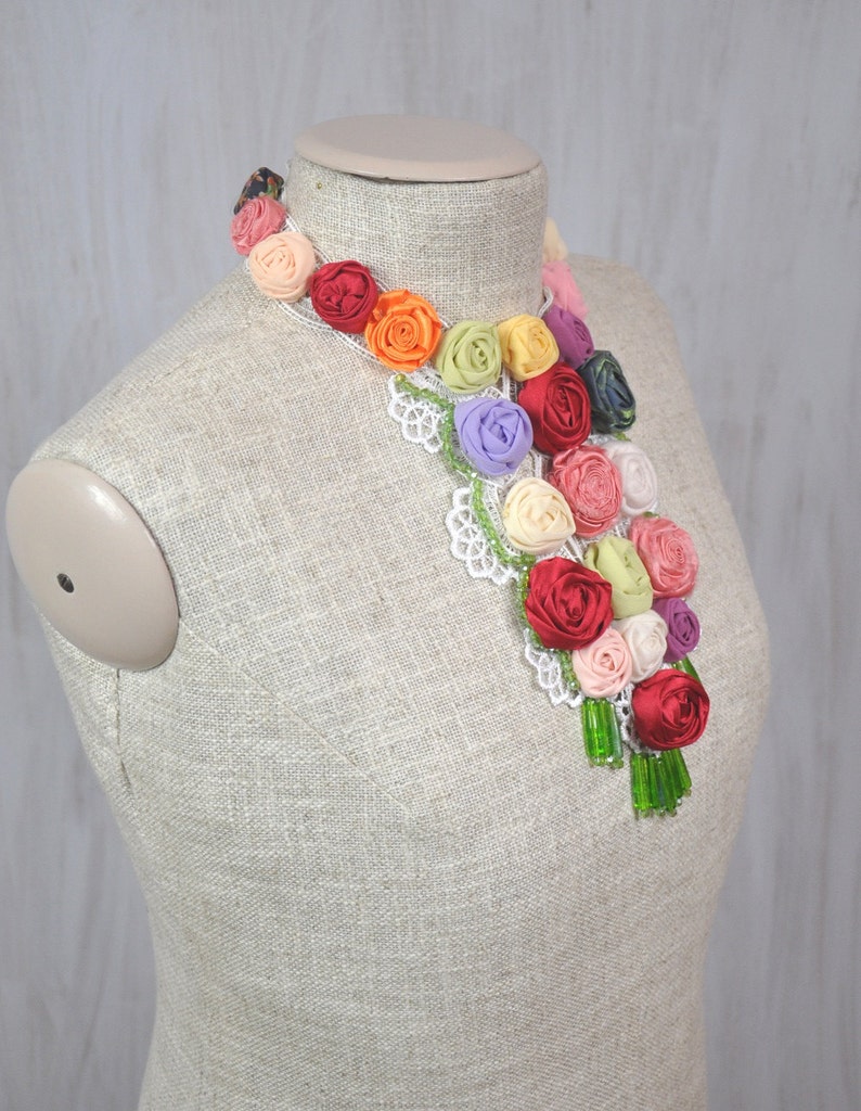 Pu&ograve; includere: Una collana floreale colorata realizzata con rosette di tessuto in tonalit&agrave; di rosso, arancio, giallo, verde, rosa e viola. La collana &egrave; decorata con pizzo bianco e perline verdi.