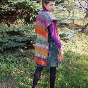 May include: A woman wearing a colorful striped crochet vest with a purple shirt and a blue denim skirt. She is standing in a grassy area with trees in the background.