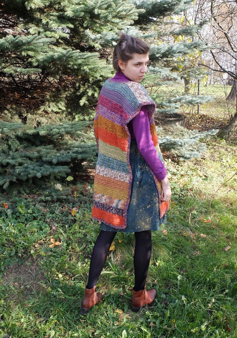 May include: A woman wearing a colorful striped crochet vest with a purple shirt and a blue denim skirt. She is standing in a grassy area with trees in the background.