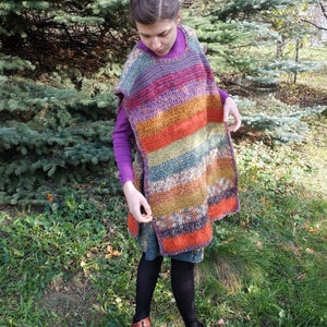 May include: A woman wearing a colorful striped crochet poncho with a purple long-sleeved shirt underneath. She is standing in a grassy area with trees in the background.