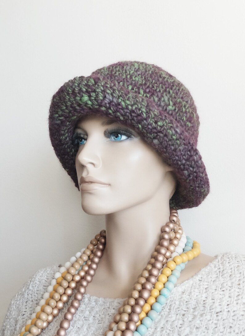 May include: A purple and green crocheted hat with a wide brim. The hat is worn by a mannequin with a multi-colored beaded necklace.