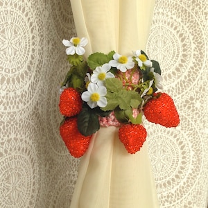 May include: A white curtain with a pink and white crocheted tie-back decorated with red strawberries and white flowers.