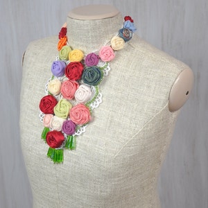 Pu&ograve; includere: Una collana floreale colorata realizzata con rose di stoffa e pizzo. La collana &egrave; esposta su un manichino beige.