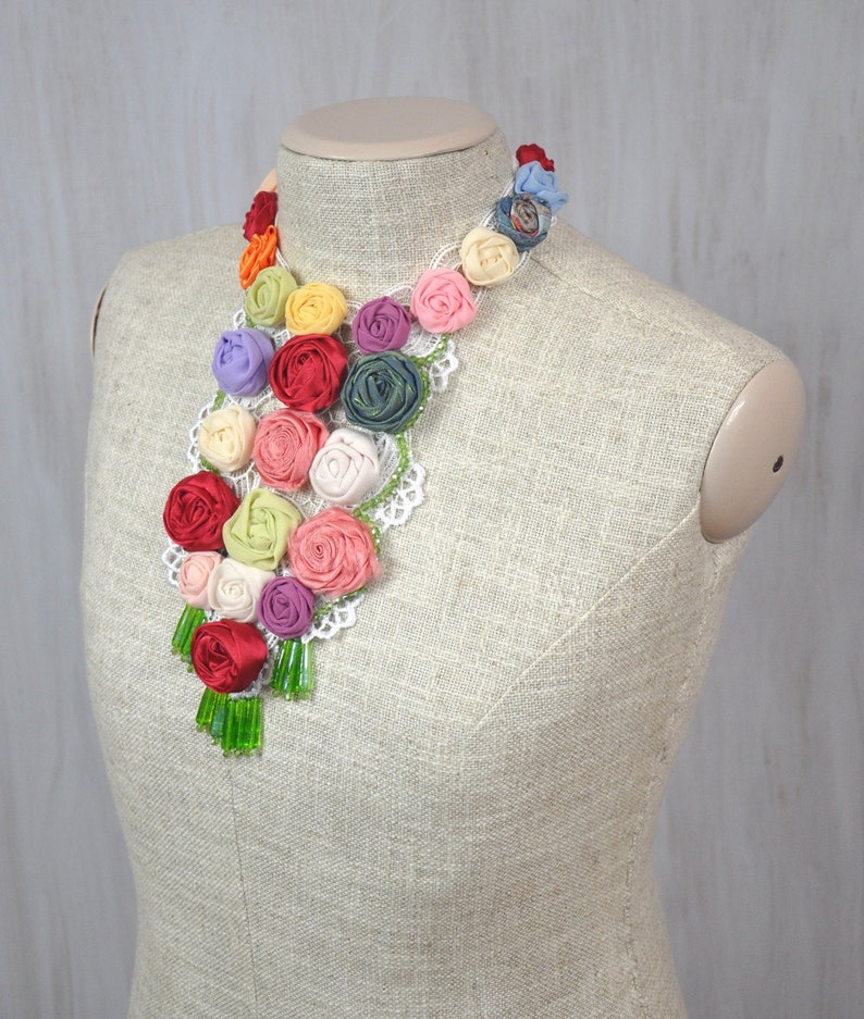 Pu&ograve; includere: Una collana floreale colorata realizzata con rose di stoffa e pizzo. La collana &egrave; esposta su un manichino beige.