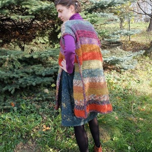 May include: A long, colorful crochet vest with a striped pattern in shades of purple, orange, brown, and green. The vest is sleeveless and has a loose fit.