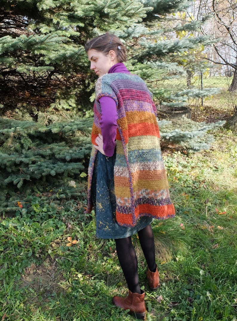 May include: A long, colorful crochet vest with a striped pattern in shades of purple, orange, brown, and green. The vest is sleeveless and has a loose fit.