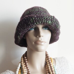 May include: A purple and green crocheted cloche hat with a wide brim. The hat is worn by a mannequin with a multi-colored beaded necklace.