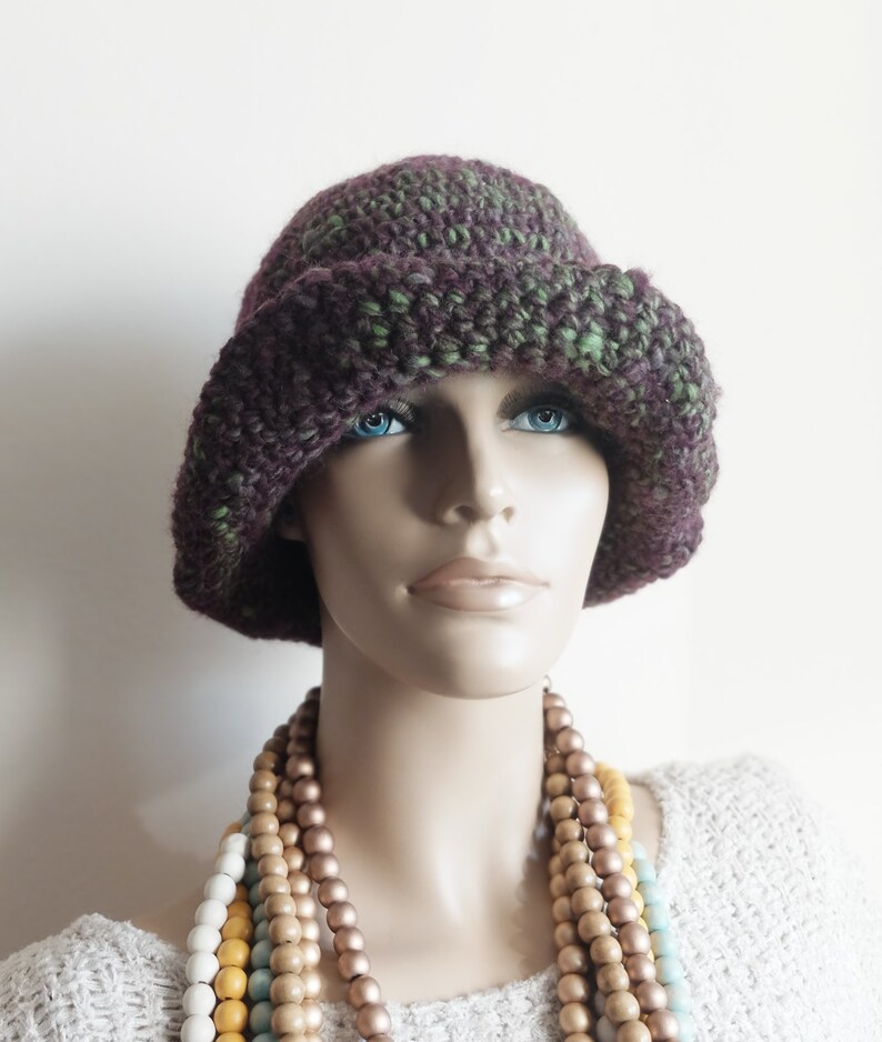 May include: A purple and green crocheted cloche hat with a wide brim. The hat is worn by a mannequin with a multi-colored beaded necklace.
