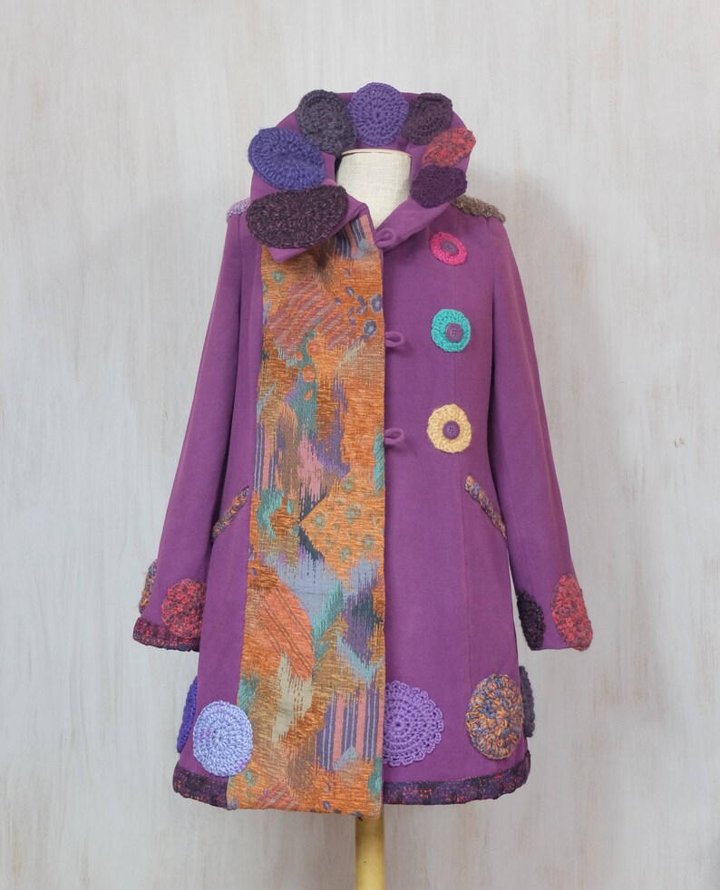 May include: A purple coat with a colorful crocheted collar and trim. The coat has a patchwork design on one side and a solid purple color on the other side.
