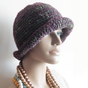 May include: A purple and green crocheted cloche hat with a wide brim. The hat is on a mannequin wearing a white shirt and a multi-colored beaded necklace.