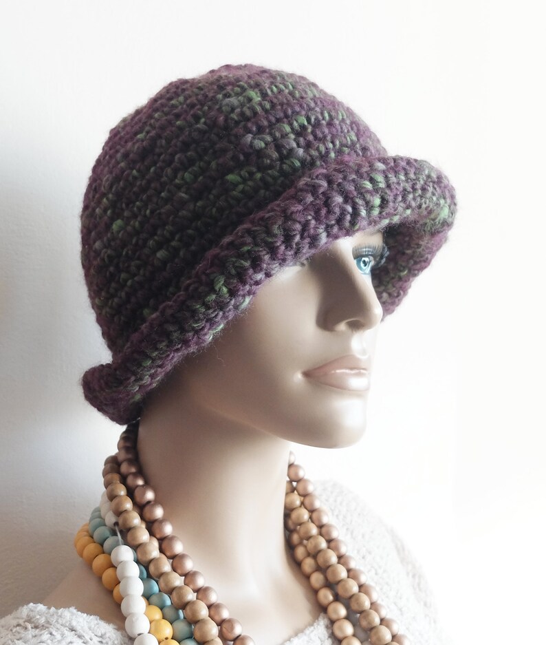May include: A purple and green crocheted cloche hat with a wide brim. The hat is on a mannequin wearing a white shirt and a multi-colored beaded necklace.