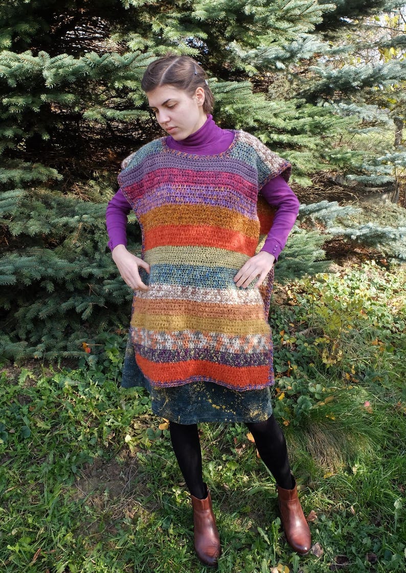 May include: A woman wearing a colorful striped crochet poncho with a purple turtleneck shirt and a dark blue skirt.