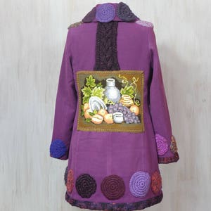May include: Purple jacket with a crocheted collar and trim. The back of the jacket features a framed embroidered image of a still life with fruit and a vase.